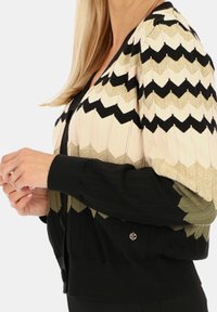 Sweter z dzianiny z wzorem w zygzak w kolorach czarnym, kremowym i oliwkowym, z ryflowanymi czarnymi mankietami oraz ozdobnym detalem w postaci guzika.