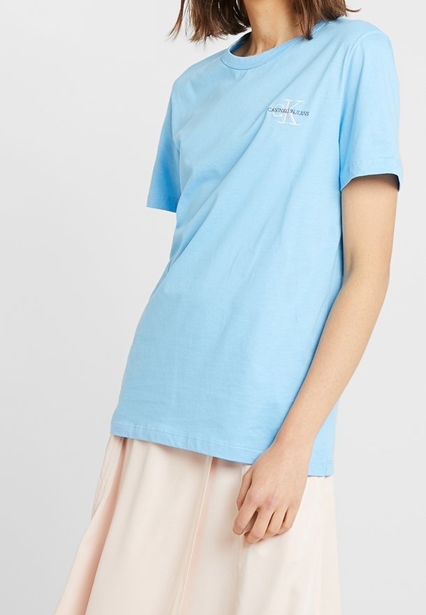 Camiseta de algodón azul claro con mangas cortas y cuello redondo. Presenta un pequeño logo en el pecho. Textura suave, ajuste casual.