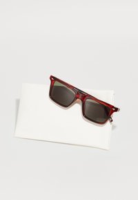 Gafas de sol de marco cuadrado con lentes oscuros, patrón de carey rojo y un estuche protector blanco con texto en relieve.