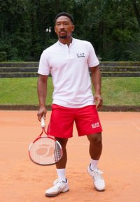 Valge lühikeste varrukatega polo, millel on lukuga kaelus ja EA7 logo, kombineeritud punaste lühikesed püksid. Mängija hoiab punast tennisereketit savimääratud väljakul.