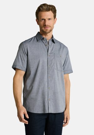 Mann trägt kurzärmliges, graues Hemd mit Brusttasche und steht vor einem einfarbigen weißen Hintergrund.