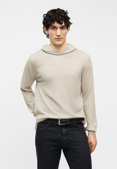 Νεαρός άνδρας με σγουρά μαλλιά, φορώντας μπεζ φούτερ με κουκούλα και μαύρα τζιν, στέκεται με το ένα χέρι πίσω από την πλάτη του.