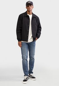 Μαύρο, ελαφρύ μπουφάν με κουμπιά με τριπλό κούμπωμα, φορεμένο πάνω από λευκό πουκάμισο. Συνδυασμένο με ανοιχτό μπλε τζιν και μαύρα αθλητικά παπούτσια με λευκές λεπτομέρειες.