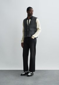 Siv volneni jopič z gumbom in dvema sprednjima žepoma, nošen preko smetanastenga rebrastega puloverja, v kombinaciji s črnimi džinsom in belimi čevlji.