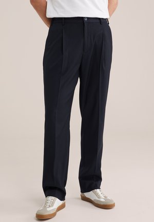 Férfi fekete rakott nadrágban és barna talpú fehér sportcipőben, egyszínű háttér előtt állva.