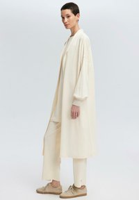 Cappotto lungo beige con maniche larghe, indossato sopra un top e pantaloni crema abbinati, abbinato a scarpe da ginnastica marroni chiare. Design minimalista.