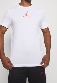 Man med vit t-shirt med korta ärmar och orange Jumpman-logotyp samt svarta shorts med matchande röd Jumpman-logotyp.