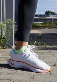 Witte sportieve schoen met oranje accenten, textuur bovenkant en geveerde zool, gecombineerd met een lichtgroene enkelsok en zwarte leggings.