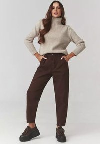 Kobieta w beżowym, prążkowanym swetrze typu golf, ciemno-brązowych spodniach z wysokim stanem i czarnych butach na platformie, stojąca z rękami w kieszeniach.