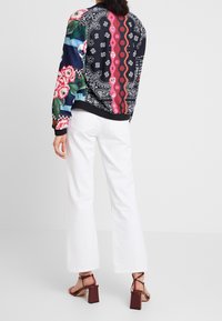 Osoba v bílých rozšířených kalhotách, vínových sandálech na podpatku a dlouhém jacketu s multibarevným vzorem obsahující florální a geometrické designy, stojí zády.