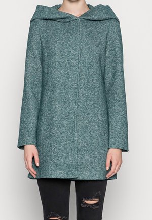 Mujer con abrigo de lana con capucha en color verde azulado y mangas largas, combinada con jeans negros rotos, posando frente a un fondo claro y sencillo.