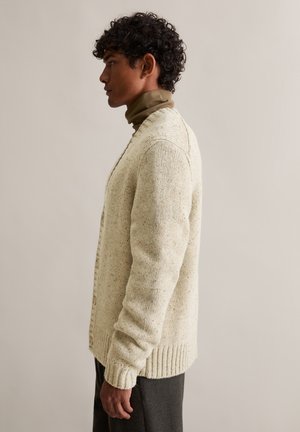 Strickjacke in hellbeige mit strukturiertem Finish, gerippten Bündchen und Knopfverschluss. Über einem dunklen Rollkragenpullover getragen.