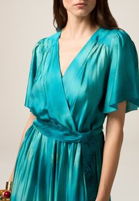 Robe portefeuille turquoise en satin fluide avec des manches courtes volantées, un décolleté en V et une taille cintrée. Présente une texture et un éclat subtils.
