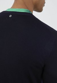 Maglione blu navy con finitura testurizzata, dotato di un bordo verde al colletto e un piccolo logo circolare nella parte posteriore del collo.