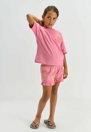 Lyserød bomulds t-shirt med korte ærmer og solgrafik, kombineret med lyserøde ternede shorts med flæsekant og grå klipklapper.