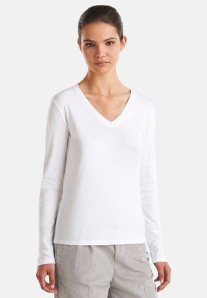 Vrouw met donker haar in een staart, gekleed in een witte langmouwen V-hals shirt en lichtgrijze corduroy broek tegen een effen achtergrond.