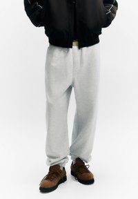 Pantaloni jogger din fleece gri cu mansete elastice si buzunare laterale, asortati cu adidasi maro texturati si o jacheta bomber neagra.