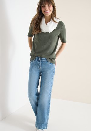 Hymyilevä nainen seisoo kädet selän takana, yllään oliivinvihreä lyhythihainen paita, valkoinen huivi ja vaaleansiniset leveälahkeiset farkut, neutraalia taustaa vasten.