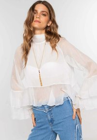 Mujer con cabello ondulado que lleva una blusa blanca de volantes transparente sobre una camiseta blanca, jeans de tiro alto azules y un largo collar dorado con borlas.