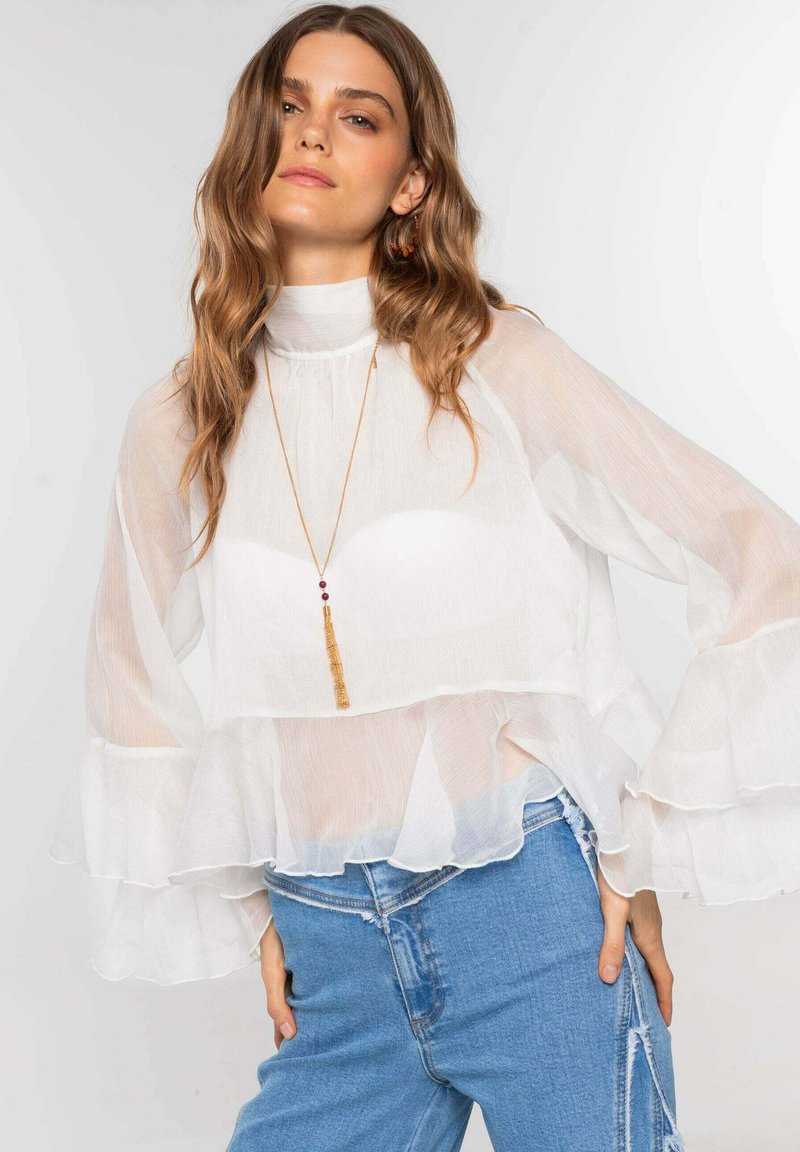 Mujer con cabello ondulado que lleva una blusa blanca de volantes transparente sobre una camiseta blanca, jeans de tiro alto azules y un largo collar dorado con borlas.