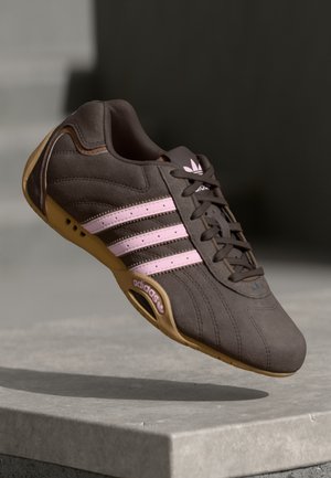 Bruine Adidas sneaker met roze strepen en logo, beige zool, zwevend boven een stenen oppervlak met een donkere achtergrond.