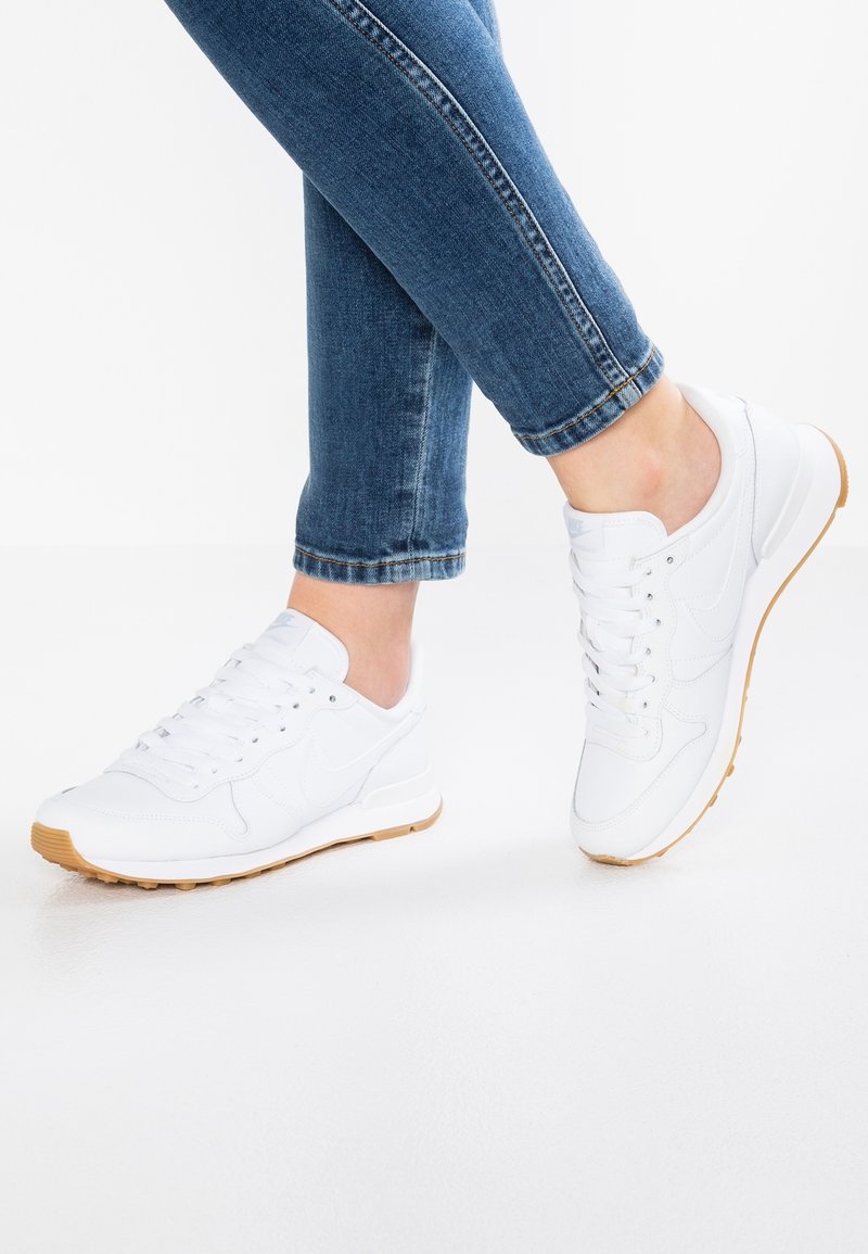 Jambes en jean bleu portant des baskets blanches à semelles en gomme, debout sur une surface blanche devant un fond blanc.