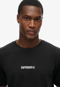 Sort bomulds T-shirt med klassisk rund hals, der har hvid broderet "SUPERDRY" logo på brystet. Glat tekstur og tætsiddende pasform.