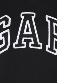 Sudadera negra con grandes letras bordadas en blanco que dicen "GAP". La textura es suave y se pueden ver detalles de costura alrededor de las letras.