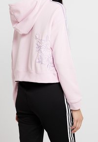 Kvinna som bär en ljusrosa kort ärmad hoodie med blommig brodyr på ryggen och svarta byxor med vita sidränder.