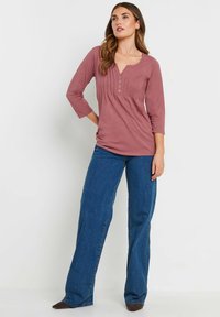 Himbeerfarbene Bluse mit 3/4-Ärmeln, Henley-Ausschnitt und vertikalen Falten. Kombiniert mit weiten indigofarbenen Jeans und dunkelbraunen Schuhen.