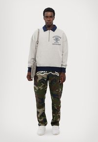 Sivá mikina s námorníckym golierom, zipsovým zapínaním a logom. Camo cargo nohavice s detailmi vrecka. Biele tenisky. Jednoduchá ramenná taška.