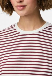 Vrouw met kort bruin haar, gekleed in een wit en kastanjebruin horizontaal gestreept T-shirt met ronde hals, te zien van kin tot borst.