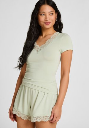 Vrouw draagt lichtgroene top met korte mouwen en bijpassende korte broek met kanten rand, staand tegen een effen achtergrond, kijkt opzij.