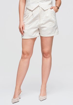 Kvinde iført hvide broderede shorts og matchende vest, parret med hvide flettede høje hæle, stående mod en ensfarvet baggrund.