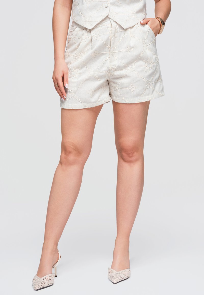 Femme portant un short blanc brodé et un gilet assorti, assortis à des chaussures à talons blancs tissées, se tenant devant un fond uni.