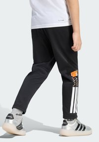 Osoba oblečená ve černých kalhotách Adidas s bílými pruhy a oranžovou grafikou, stříbrných teniskách Adidas a šedých ponožkách Adidas, stojící na světlém pozadí.