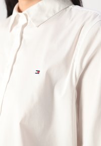 Witte overhemd met kraag, gemaakt van een gladde stof, met een klein geborduurd logo in rood, wit en blauw op de linkerborst.