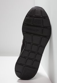 Svart Adidas sneaker sula med texturerade gummigreppmönster och Adidas-logotyp i mitten, vinklad mot en vit yta.