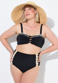 Černý bikiny set s otočeným předním designem, bílým vlnkovaným lemováním a vysokými spodními kalhotkami s mřížkovým detailem. Viditelný slaměný klobouk.