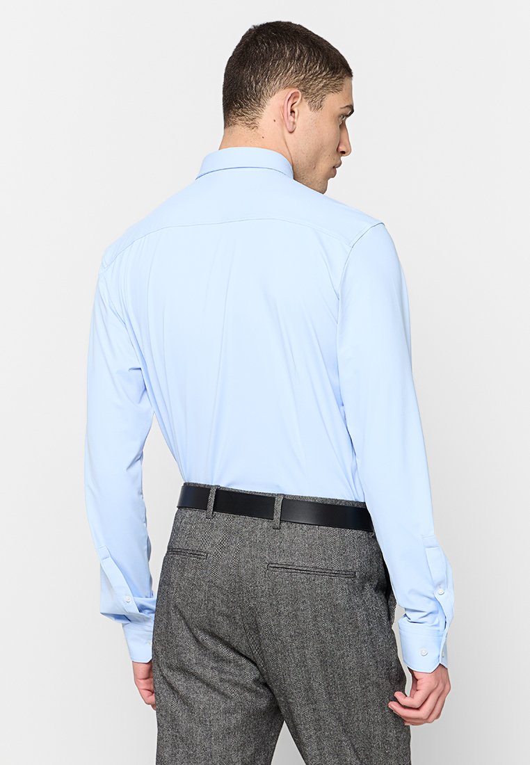 Chemise bleu clair à manches longues avec un empiècement au dos, coupe ajustée et poignets boutonnés, assortie à un pantalon gris texturé et une ceinture noire.