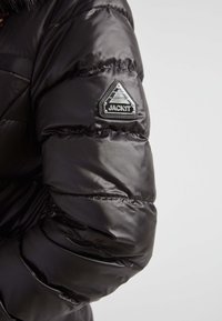 Czarna kurtka puchowa o błyszczącej powierzchni, z pikowanymi sekcjami. Na lewym rękawie znajduje się trójkątny naszywka z tkaniny z logo "JACK1T".