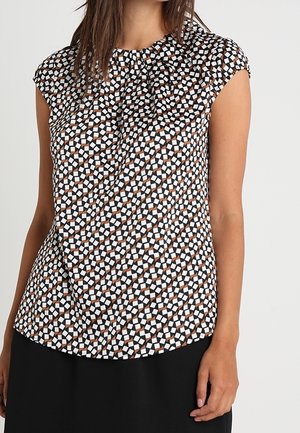 Femme portant un chemisier sans manches avec un motif géométrique noir, blanc et marron et un pantalon noir sur un fond uni.