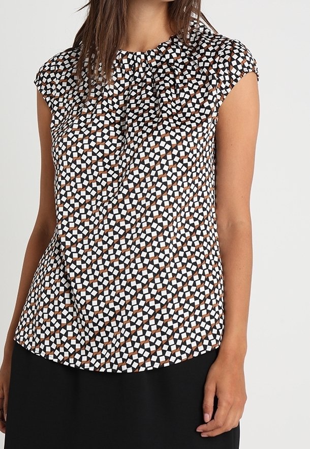 Femme portant un chemisier sans manches avec un motif géométrique noir, blanc et marron et un pantalon noir sur un fond uni.