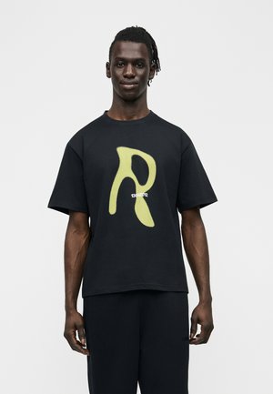 Mężczyzna z zaplecionymi włosami stoi przodem, ubrany w czarną koszulkę z dużą żółtą stylizowaną literą "R" i małym białym napisem.