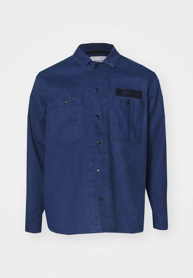 Jeansowa koszula w ciemnoniebieskim kolorze z długimi rękawami, spiczastym kołnierzykiem, dwiema kieszeniami na piersi oraz czarną naszywką z logo po lewej stronie na górze.