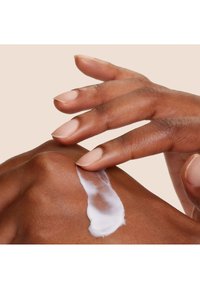 Une lotion blanche appliquée sur une peau marron, avec une main qui l'étale doucement. La main a des ongles naturels et une texture lisse. Fond doux.
