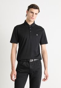 Black polo shirt with short sleeves, three-button placket, and a small round logo on the left chest, paired with black jeans.