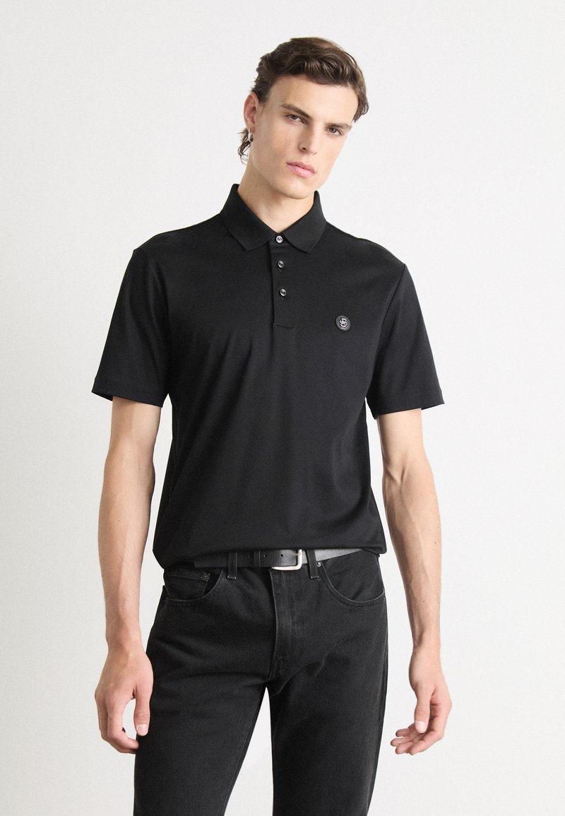 Black polo shirt with short sleeves, three-button placket, and a small round logo on the left chest, paired with black jeans.