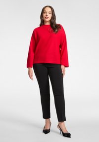 Roter Pullover mit langen Ärmeln in lockerer Passform, kombiniert mit schwarzen, schmal geschnittenen Hosen und schwarzen High-Heels mit Schleifenakzenten. Glatte Texturen.