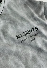 Pilkas medvilninis marškinėlis su subtiliu tekstūros raštu. Turi apvalų kaklą ir juodai atspausdintą tekstą: "ALLSAINTS," su papildomais simboliais ir "LONDON."